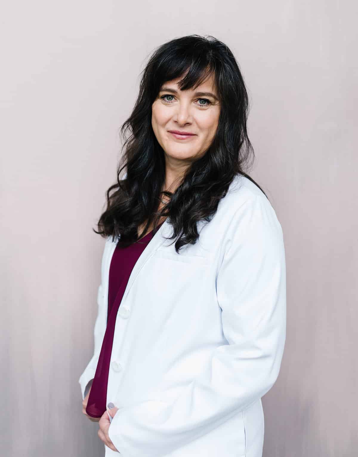 A woman with dark hair wearing a white coat and a maroon top, specializing in pregnancy care, standing against a light pink background.