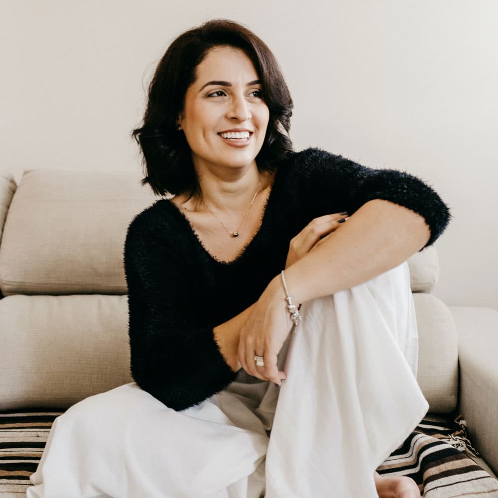 A smiling woman in a black sweater and white pants sitting on the floor leaning against a couch.