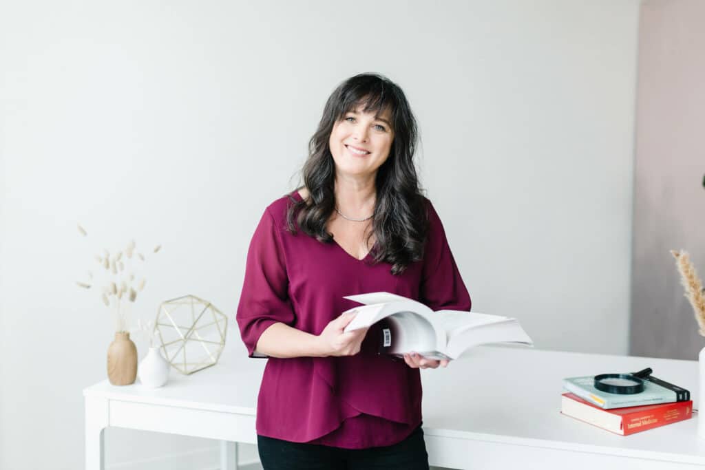 A woman in a magenta blouse smiling and holding an open book about fertility acupuncture in a light, modern room.