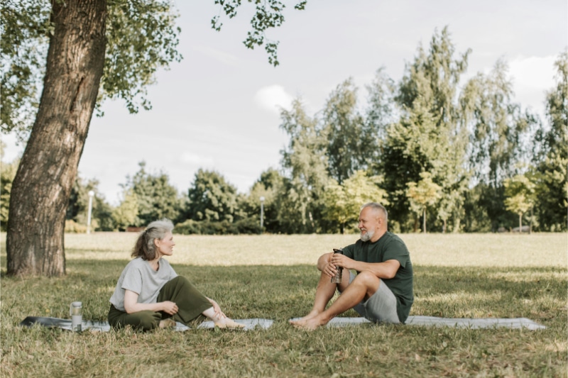 Older man and woman sit on yoga mats under a tree as they discuss what causes sciatic nerve pain.