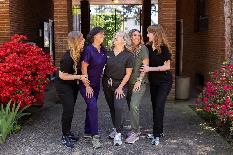 The Willow Tree Clinic team laughing together outside the clinic, standing near vibrant red azaleas and a brick archway.