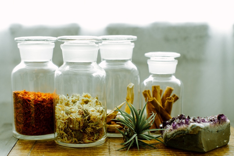 Four glass apothecary jars filled with colorful dried herbs like saffron, chamomile, and cinnamon sticks sit on a wooden table next to a purple crystal.