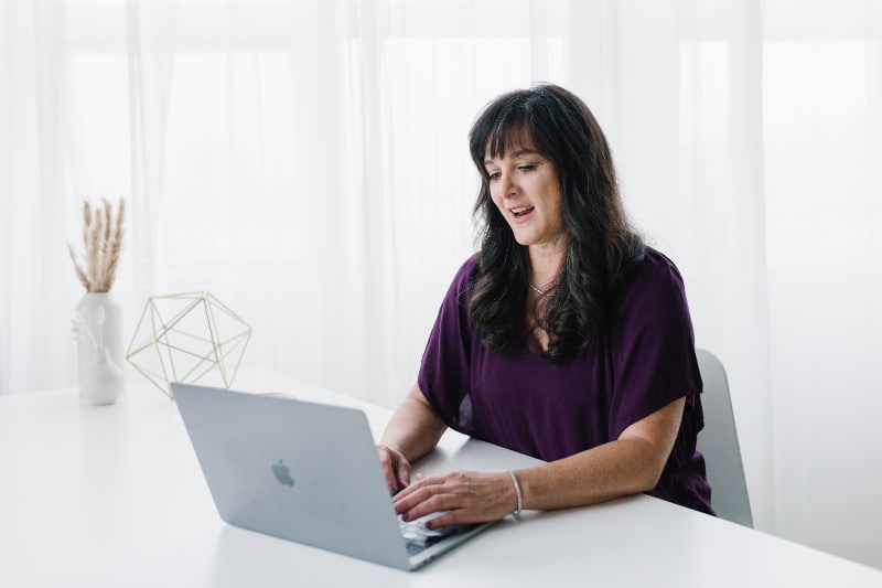 Woman in purple top working on a laptop at a white desk, representing virtual fertility consultation or health coaching. | acupuncture vs ivf