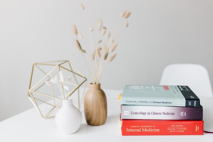 Minimal white desk with a geometric gold decor piece, two small vases with dried grasses, and a stack of Chinese medicine and gynecology textbooks.